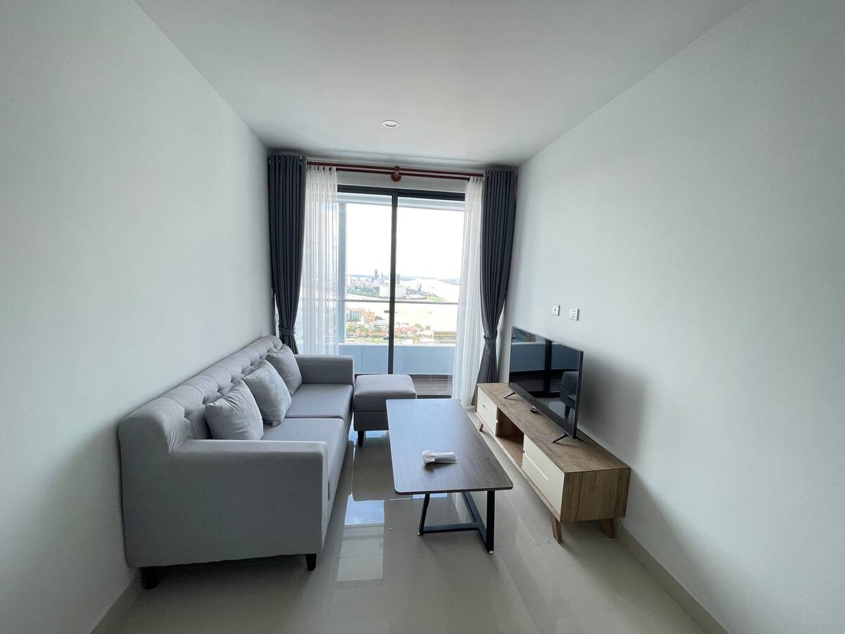 A bright living area features a comfortable grey sofa positioned next to a wooden coffee table. Large windows provide natural light and showcase views of the city and river. A television is mounted on the opposite wall, enhancing the space's functionality.