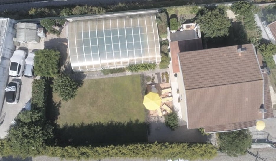 An aerial view captures a spacious lawn surrounded by hedges and greenery. A large, clear greenhouse is visible, along with a sun patio featuring a yellow umbrella. The house displays a brown roof, and several vehicles are parked along the perimeter.