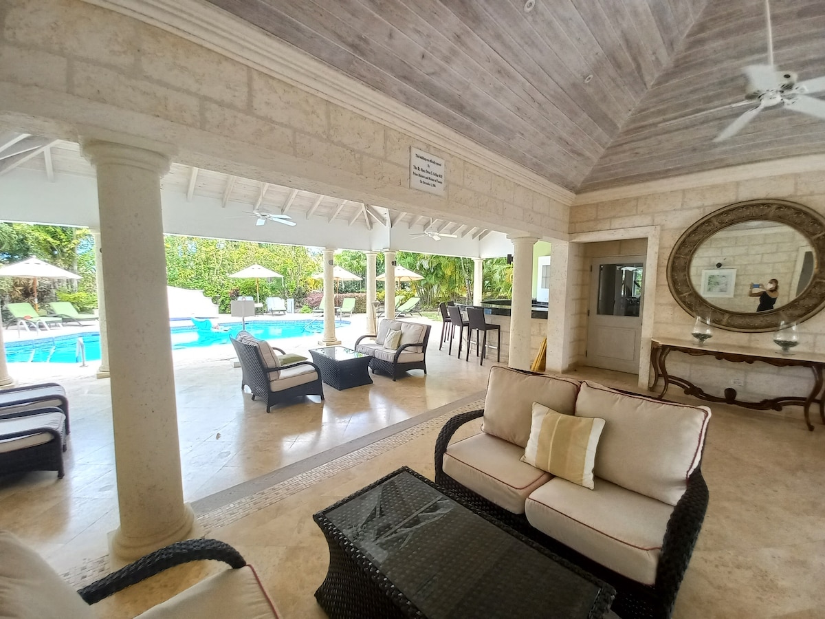 An open-air seating area is featured, with comfortable wicker furniture arranged around a glass coffee table. Light floods in from the large window overlooking the swimming pool, which is surrounded by lush greenery and shaded loungers. Ceiling fans enhance the cooling atmosphere.