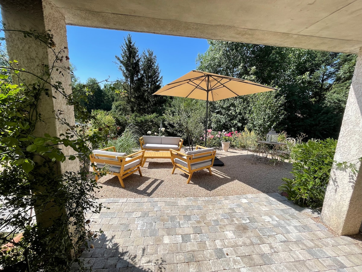 An outdoor seating area is visible under a large umbrella, surrounded by lush greenery and colorful flowers. Two lounge chairs and a sofa are arranged on a gravel surface, with a small table nearby, creating a serene space for relaxation.