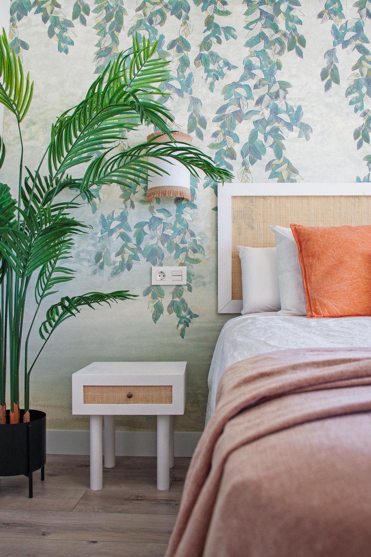 A serene bedroom corner is highlighted by a stylish bed adorned with soft, neutral bedding and an accent pillow. A decorative plant stands beside a bedside table featuring a woven design. A wall adorned with leafy wallpaper creates a calming backdrop.