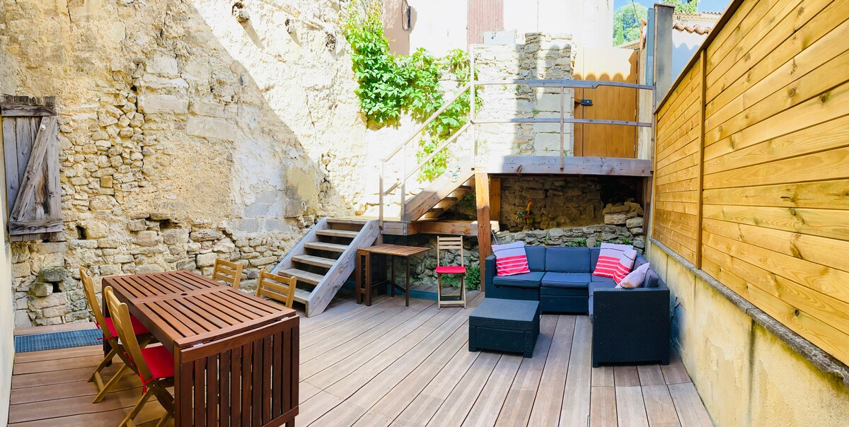 An outdoor terrace features wooden decking and stone walls, creating a relaxed atmosphere. A dining table with multiple chairs is present, alongside a comfortable seating area with cushions. Steps lead to an elevated section, enhancing the space's charm and functionality.
