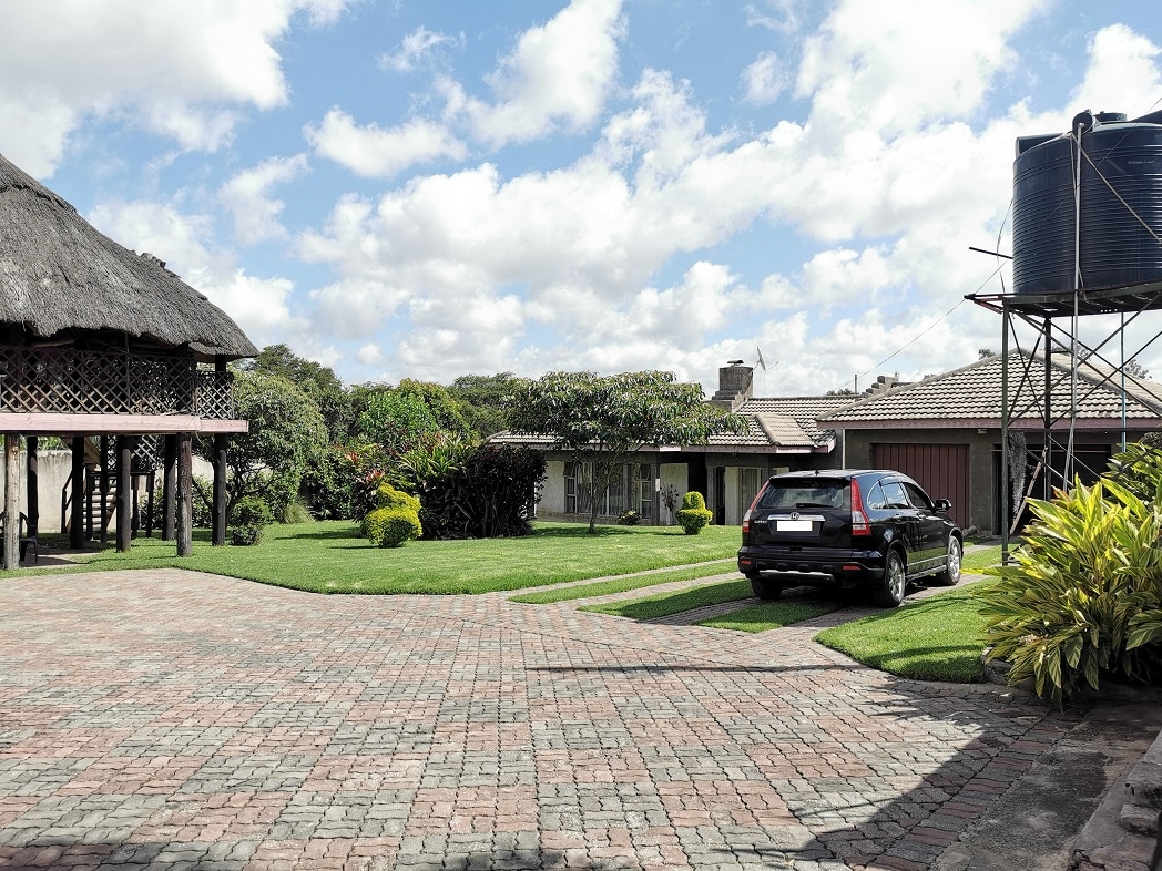 The exterior view showcases a well-maintained yard with neatly trimmed grass and vibrant shrubs. A driveway is visible with a parked vehicle, and a tall water tank stands near a gazebo. Additionally, a series of clouds can be seen against a bright blue sky.