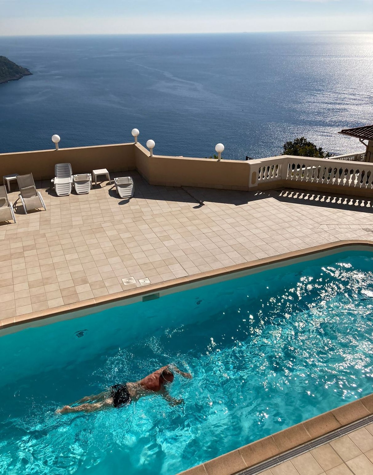A swimming pool is visible, surrounded by a tiled deck with several lounge chairs positioned nearby. The clear water reflects the sunlight, and the expansive view of the Mediterranean Sea is seen in the background, emphasizing a serene atmosphere.