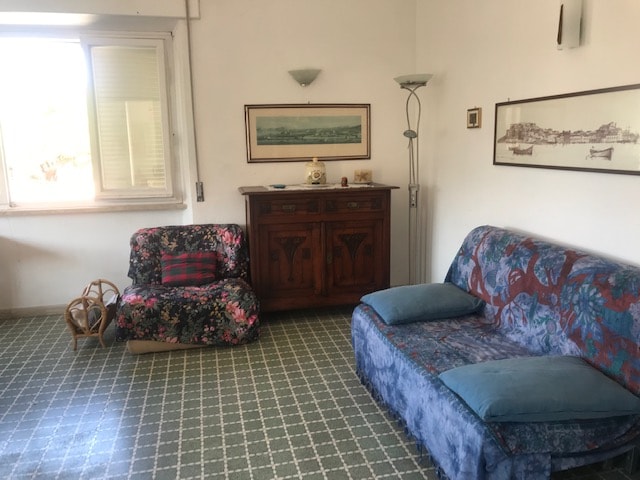 The spacious living area features a floral-patterned armchair alongside a blue sofa. A wooden sideboard is placed against the wall, while framed art and a standing lamp provide a decorative touch. Natural light enters through a window, illuminating the patterned green tile flooring.