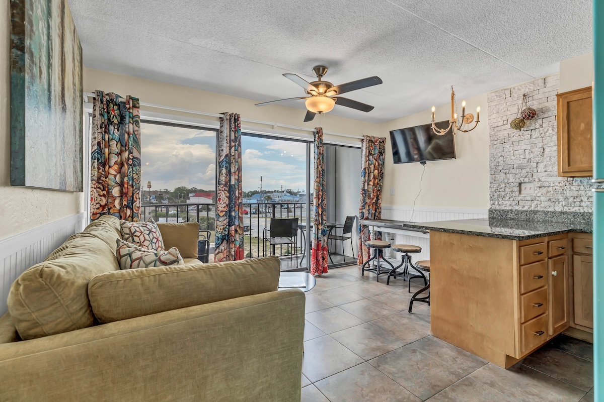 A cozy living space features a comfortable sofa and a ceiling fan. Large windows are adorned with colorful curtains, allowing natural light to fill the area. A dining table and chairs are positioned nearby, with granite countertops visible in the kitchen area.