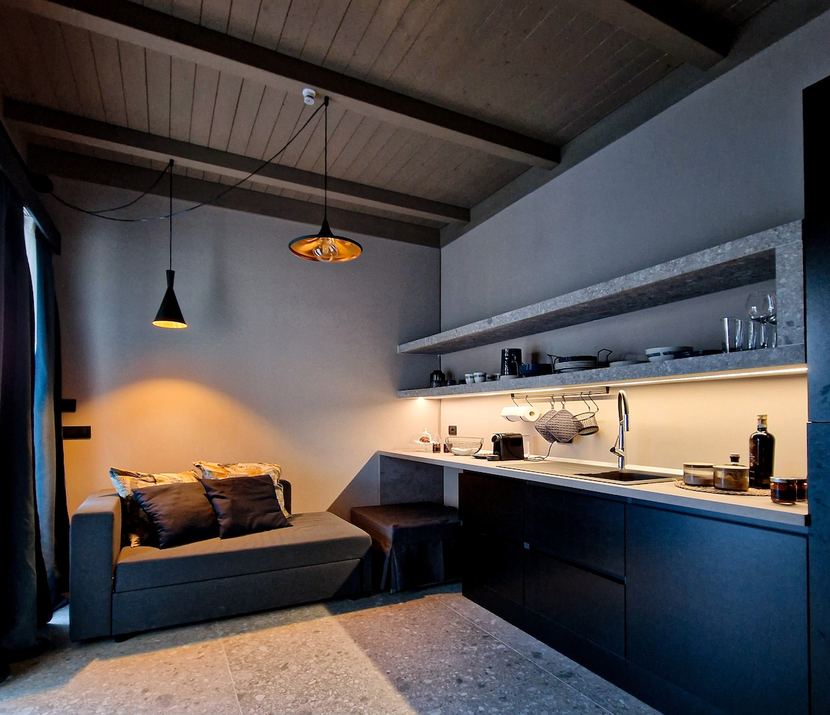 The living area features a modern sofa with decorative pillows, complemented by a sleek kitchenette with under-cabinet lighting. A shelf holds dishware and glasses, while a focused light fixture illuminates the space. Neutral tones create a calm and elegant environment.