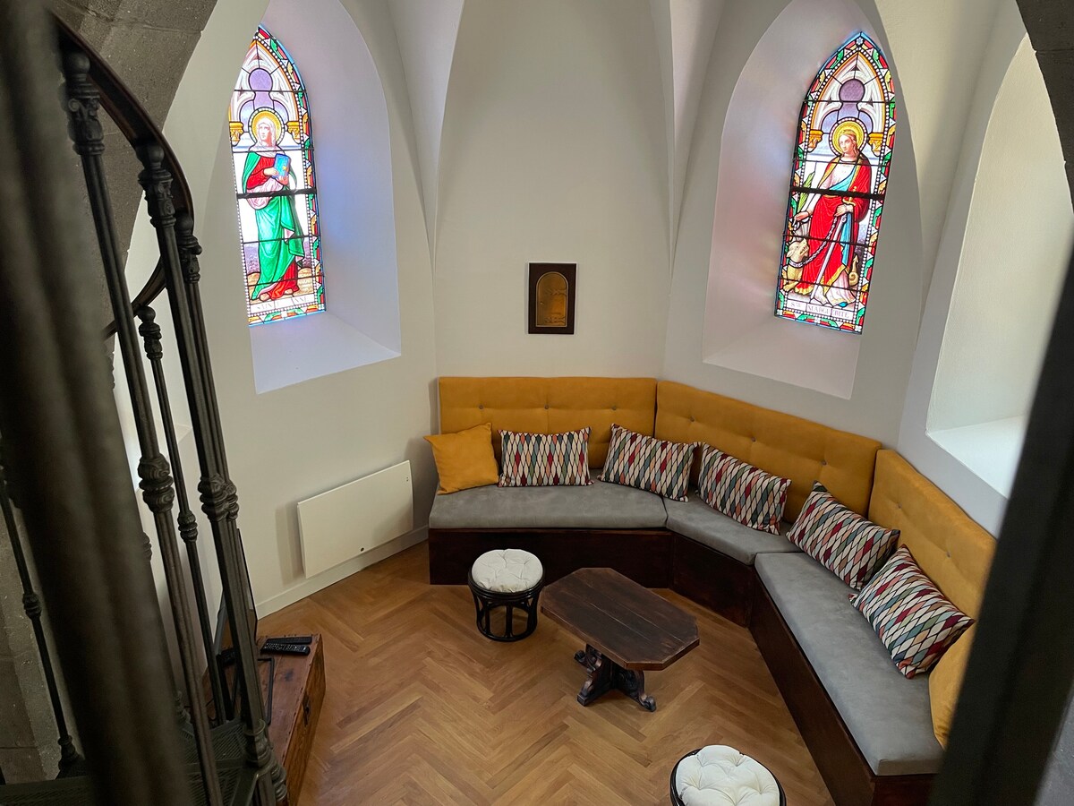 A cozy seating area features a curved, plush couch adorned with patterned cushions. A wooden coffee table occupies the center, complemented by two small stools. Bright stained glass windows illuminate the space, adding character to the room with historical touches.