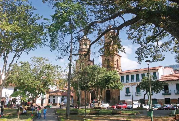 A lively square features two tall church towers framed by lush green trees. People can be seen enjoying the space, while cars line the street nearby. The surrounding buildings showcase traditional architecture and large windows, contributing to the welcoming atmosphere.