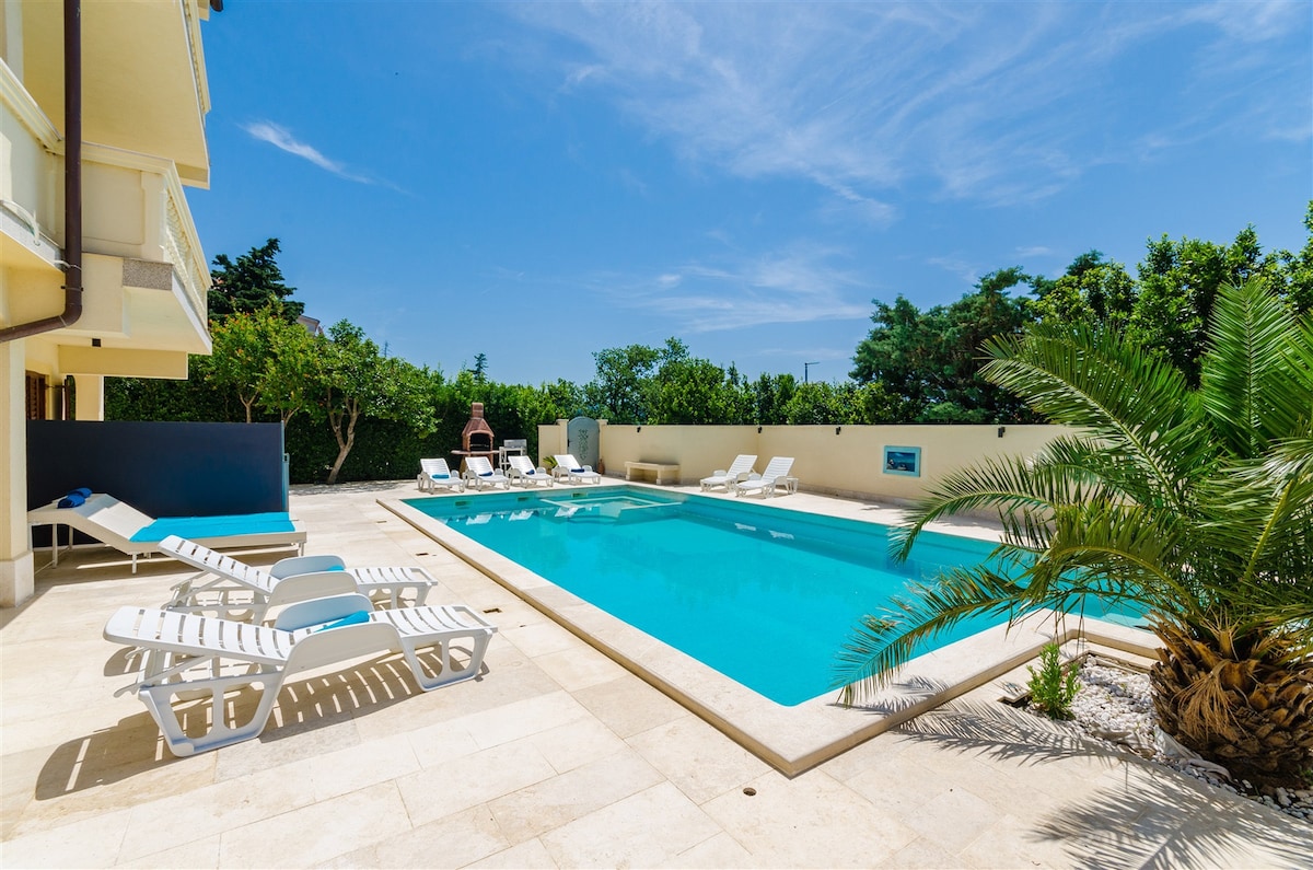 A large outdoor pool area is surrounded by a spacious deck featuring several lounge chairs. Lush greenery and palm trees provide a tranquil setting. Clear blue skies above enhance the inviting atmosphere, complemented by the pool's crisp water reflecting sunlight.