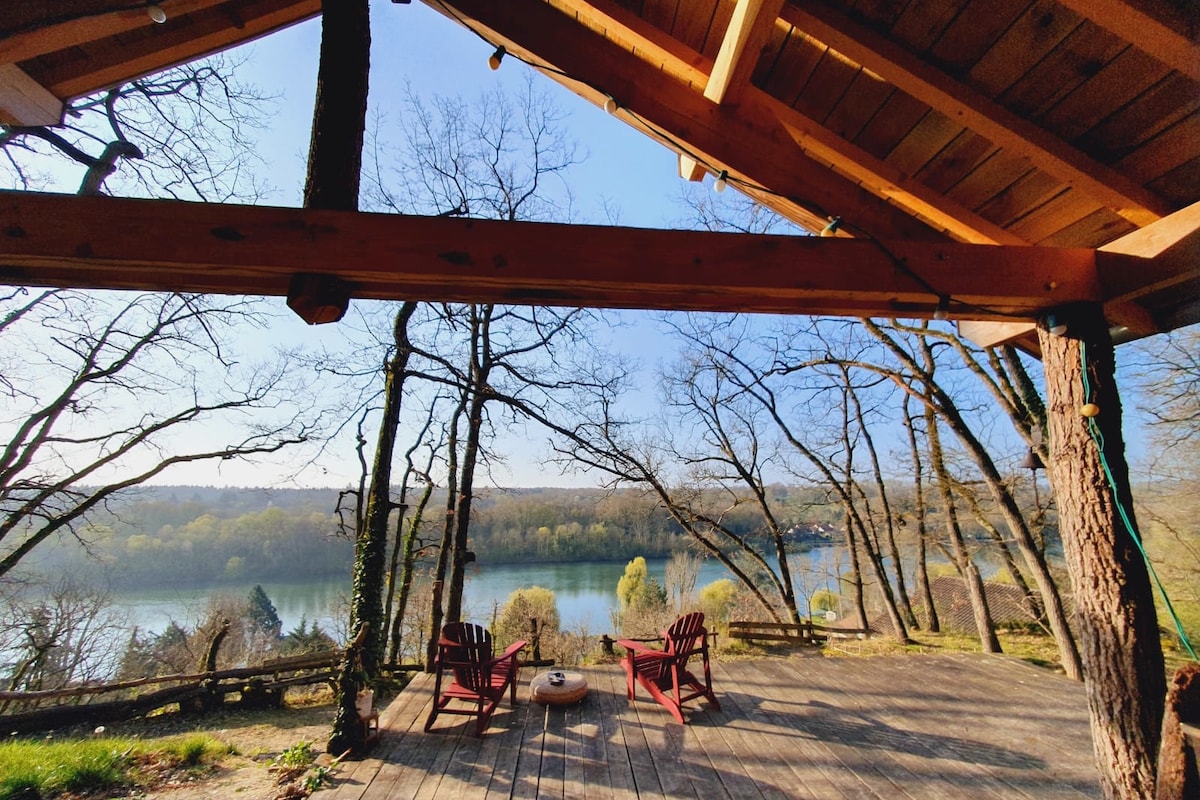 A spacious wooden terrace features two Adirondack chairs surrounding a small table, offering a view of the serene river framed by tall trees. Natural light illuminates the area, enhancing the connection to nature.