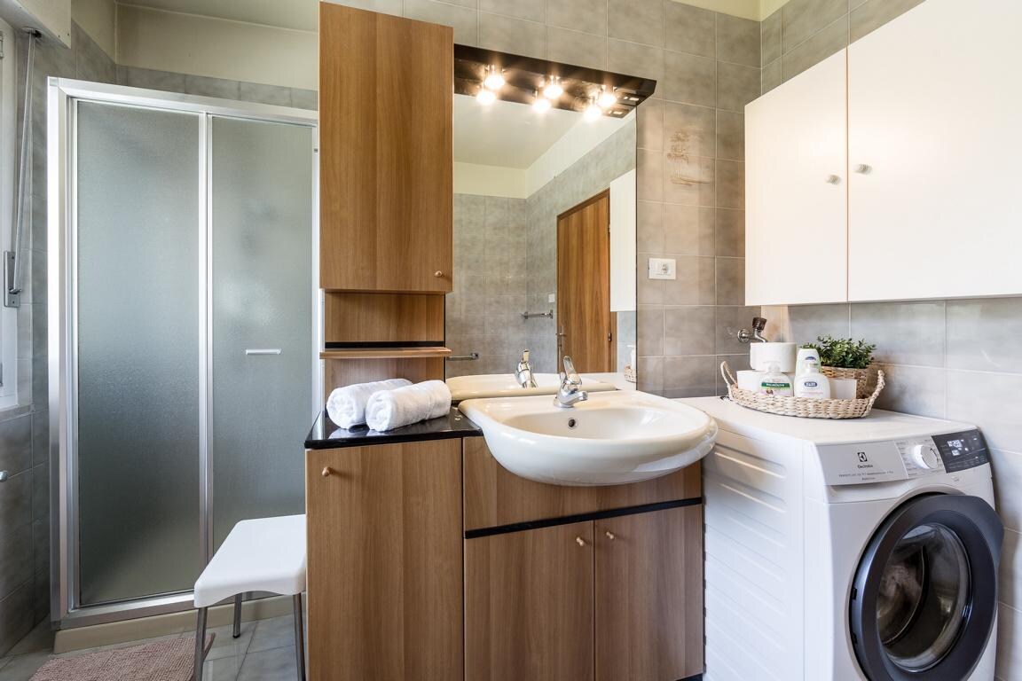 A modern bathroom features a glass shower enclosure and sleek cabinetry in warm wood tones. A round sink is mounted on a black countertop, while a washing machine is positioned beside it. Soft towels are neatly arranged on a chair, adding to the organized layout.
