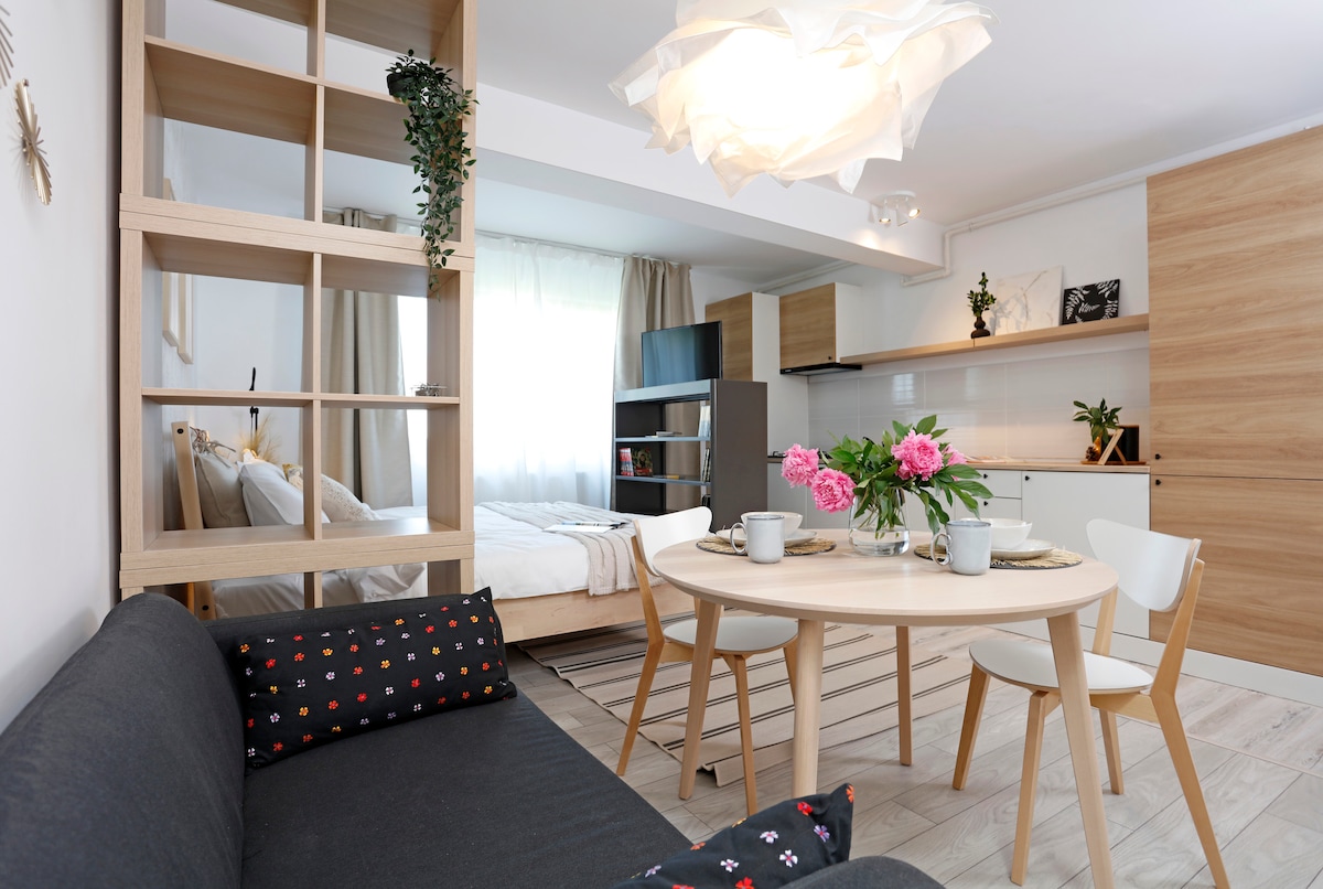 A modern studio space is depicted, featuring a comfortable green-grey couch with decorative pillows. A wooden table is set with two cups and a floral centerpiece. In the background, a cozy sleeping area with light curtains and a well-equipped kitchen can be seen, illuminated by natural light.