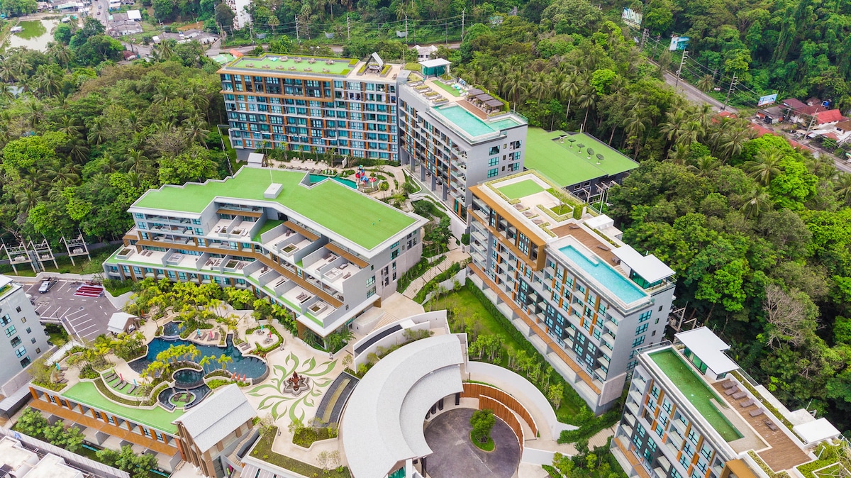 An aerial view showcases a contemporary condo complex surrounded by lush greenery. Multiple buildings with green rooftops feature outdoor pools and terraces. The landscaped courtyard includes a pool and pathways, blending seamlessly with the natural environment.