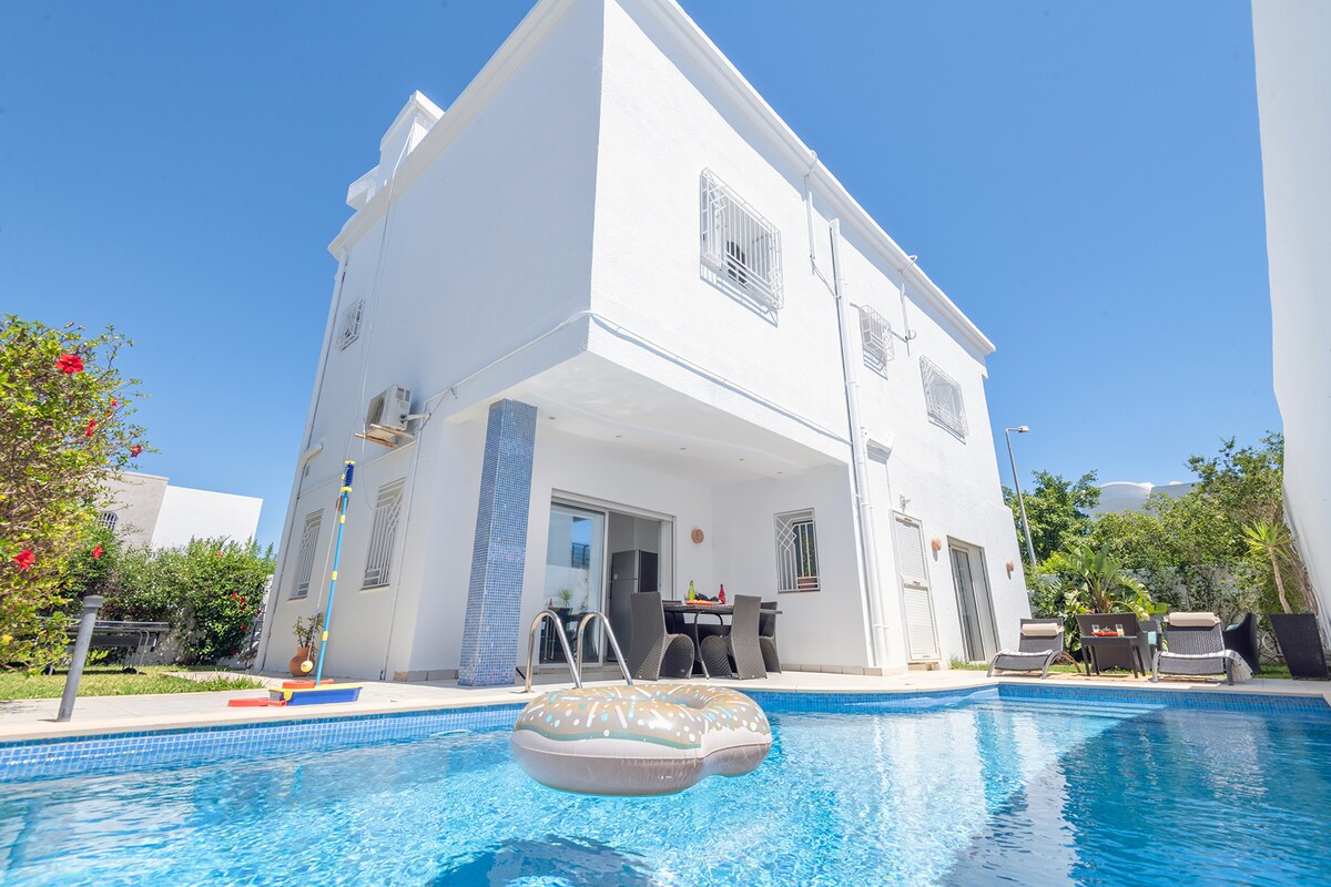 A spacious white villa is showcased, featuring large windows and a terrace overlooking a blue swimming pool. A floating inflatable ring is positioned in the pool, while surrounding greenery adds a natural touch. Outdoor seating arrangements are visible beside the water.