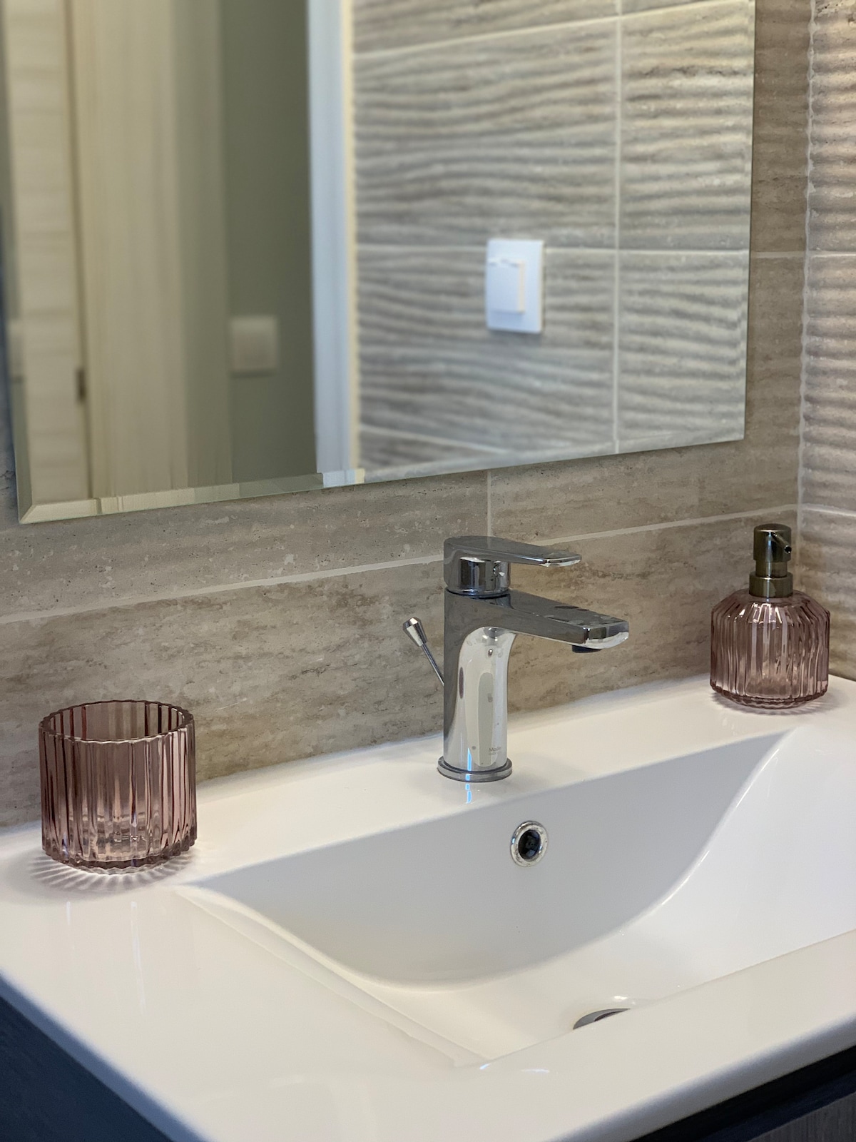 A modern bathroom sink features a sleek chrome faucet situated above a clean white countertop. Two textured glass vases in soft pink are placed on either side of the sink, complementing the neutral tile backdrop and enhancing the serene environment.