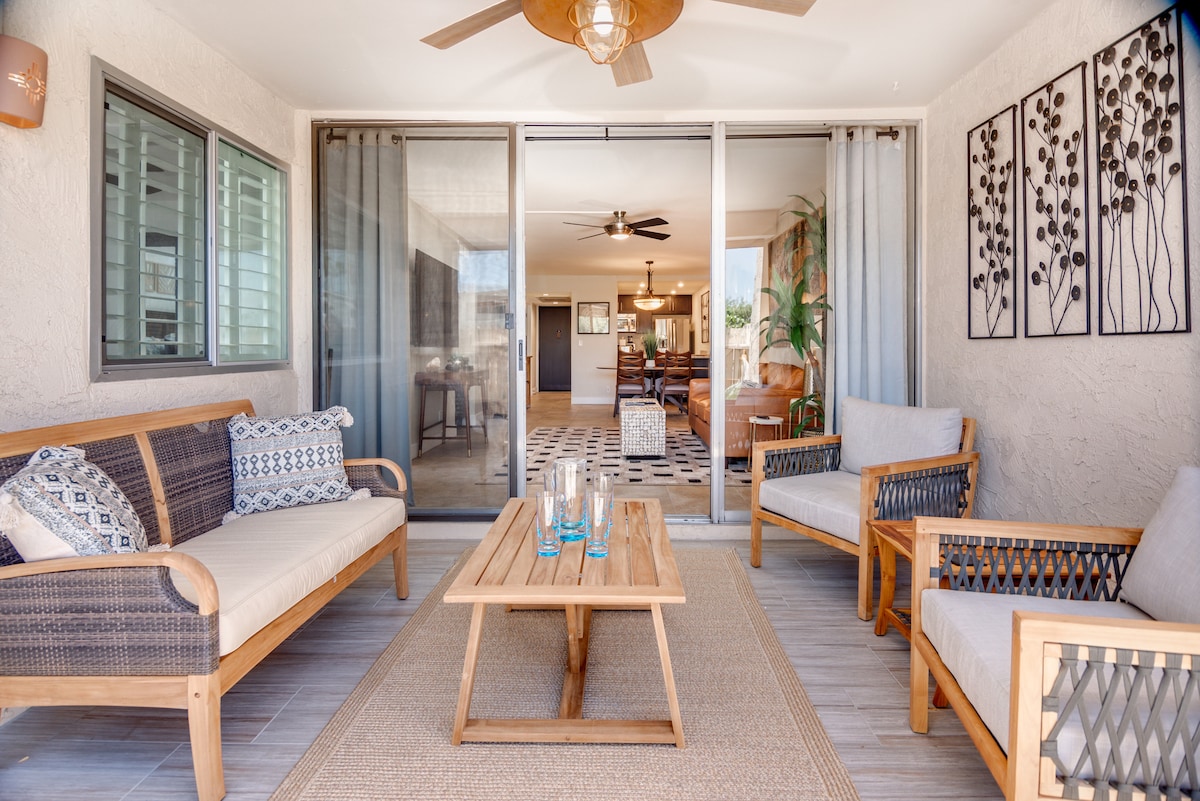 A cozy patio area is showcased with a wooden coffee table positioned between two comfortable seating groups. Natural light filters through sliding glass doors, framing a glimpse of the interior living space. Decorative wall art adds a subtle touch to the relaxing environment.