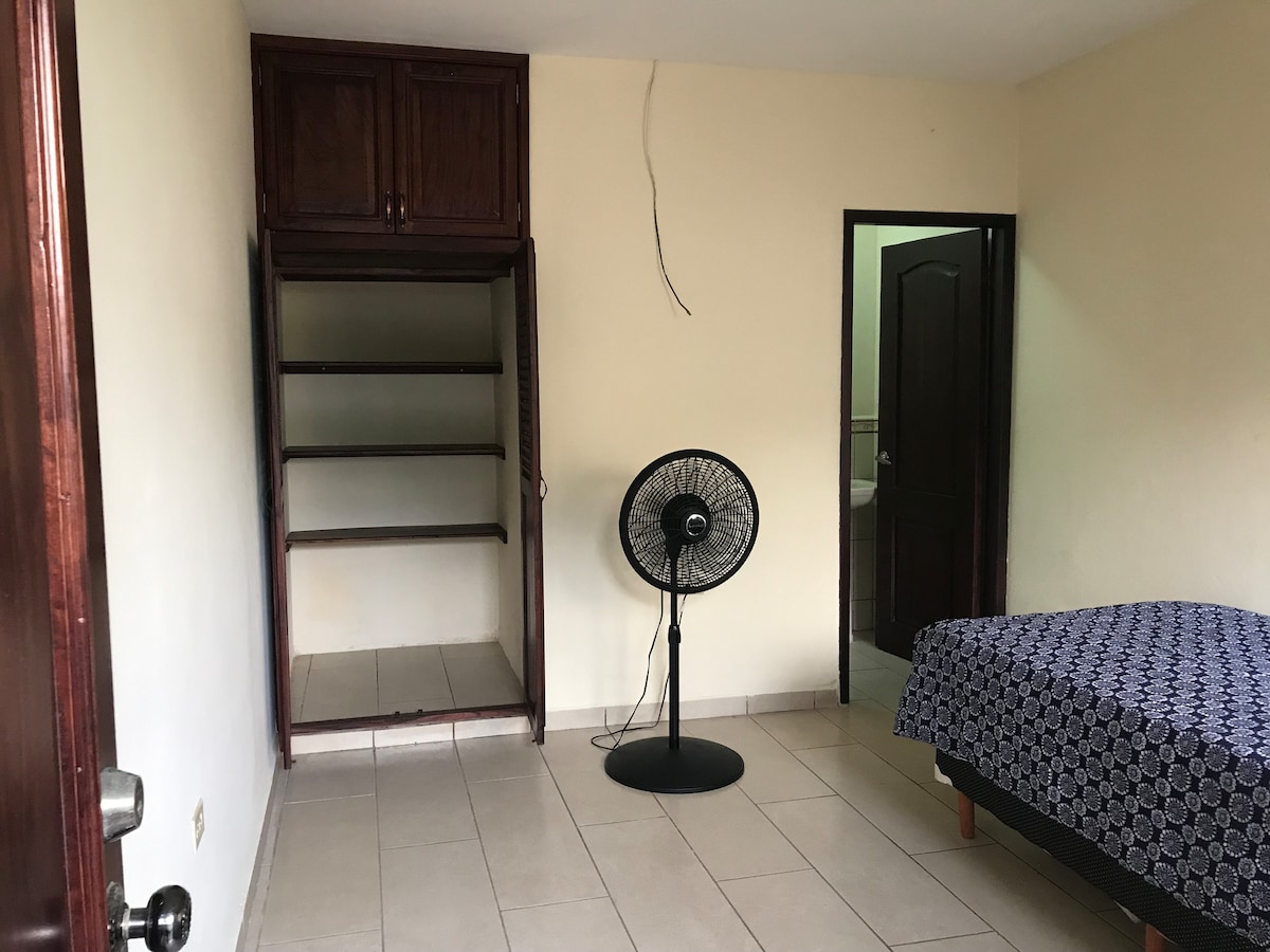 A simple room features a spacious bed covered with a patterned blue bedspread. A small standing fan provides airflow. A closet with open shelves is visible along one wall, while a doorway leads to a separate area, suggesting additional space and privacy.