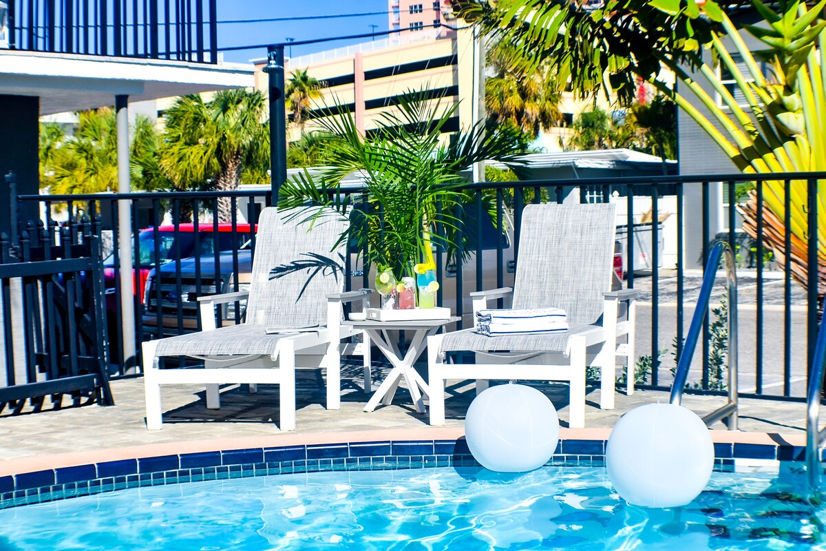 Two white lounge chairs are positioned beside a sparkling pool, accompanied by a small table adorned with colorful drinks. Lush green plants provide a touch of nature, while round white lights add a modern element to the inviting outdoor space.