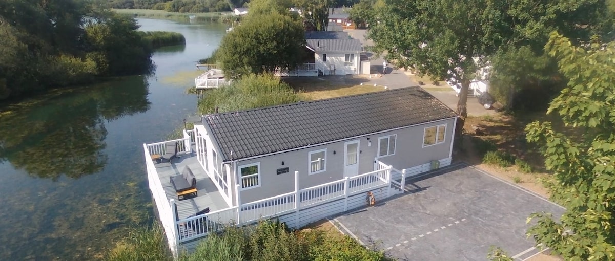 A modern lodge is positioned by a serene lake, offering an expansive outdoor decking area with seating. The exterior features light-colored walls complemented by a dark roof. A parking area for two cars is visible adjacent to the lodge, surrounded by greenery.