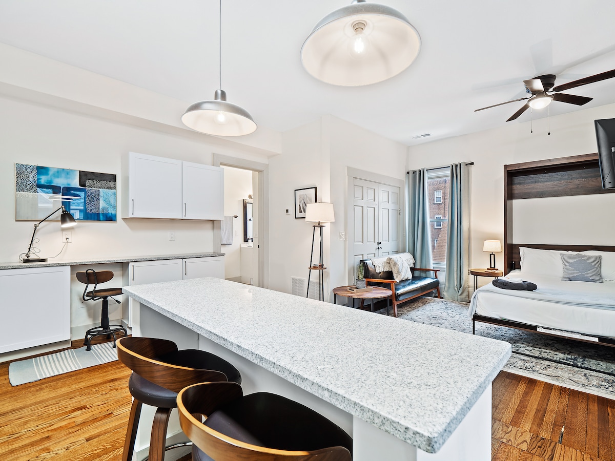 A well-lit studio space is depicted, featuring a kitchen island with three wooden stools. A comfortable seating area is seen, accompanied by a bed with neutral-toned linens. Light fixtures hang from the ceiling, contributing to the bright atmosphere. Large windows provide natural light.