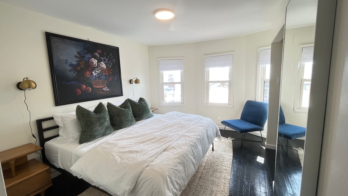 A well-lit bedroom features a spacious bed adorned with decorative pillows. A large mirror reflects the room, which includes a cozy blue chair and a striking floral artwork on the wall. Natural light streams in through three windows, enhancing the inviting atmosphere.