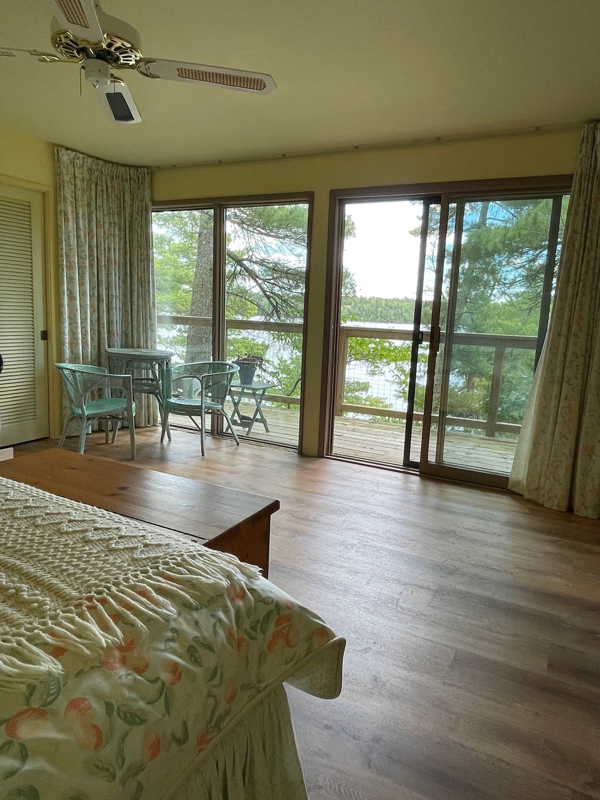 A serene bedroom area features a spacious bed adorned with a patterned quilt. Large sliding glass doors open to a private deck with a table and two chairs, offering views of the surrounding trees and water. Natural light fills the room through the curtains.