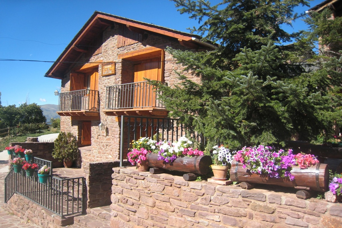 The restored farmhouse displays rustic stonework and wooden accents, featuring two balconies adorned with wooden railings. Colorful flower pots with vibrant blooms line the stone wall, creating an inviting entrance. Green trees enhance the natural surroundings, with a clear blue sky overhead.