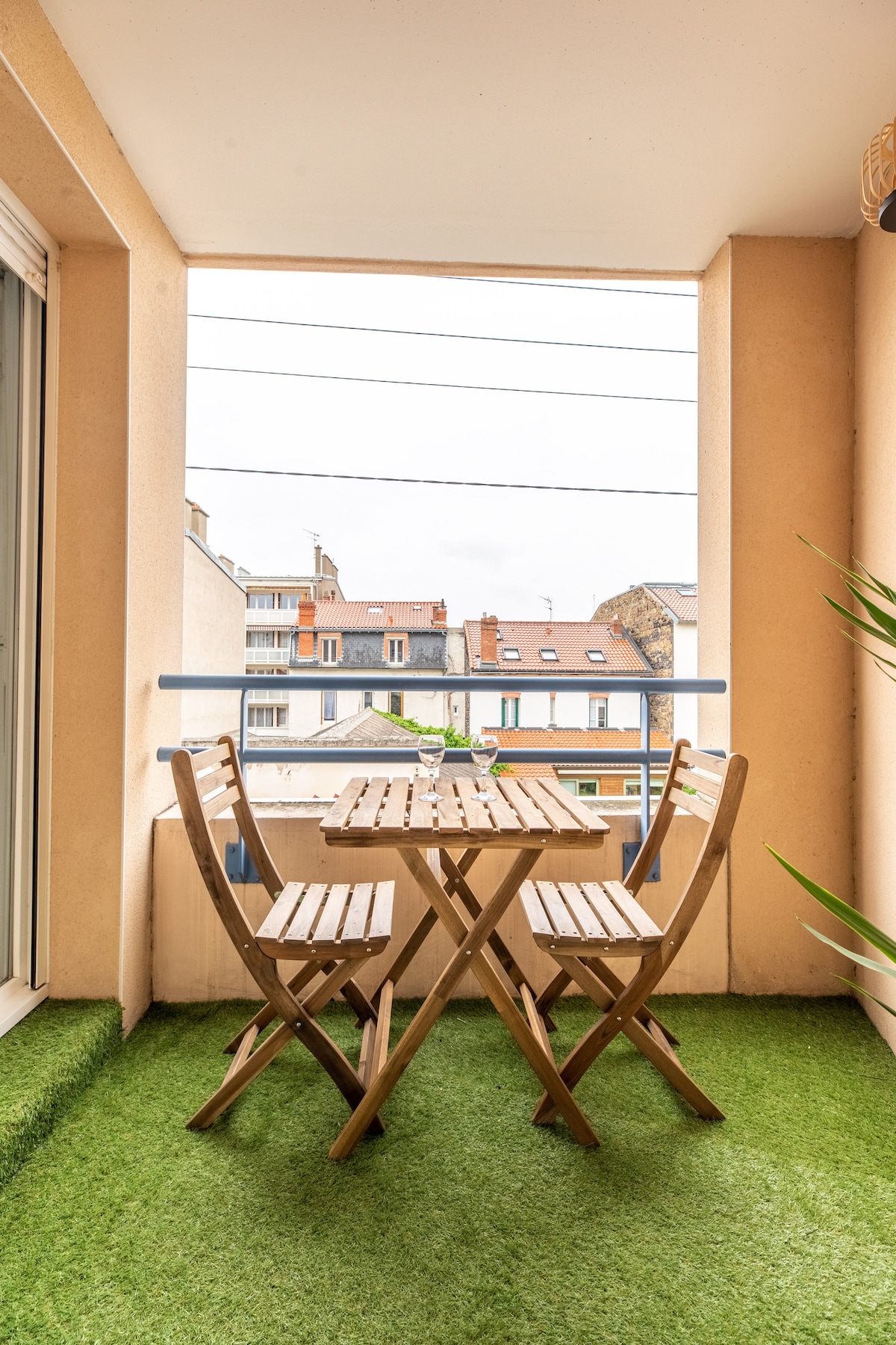 A small terrace is presented with a wooden table and two folding chairs, situated on green artificial grass. Natural light is captured through surrounding windows, offering a view of the neighboring buildings.