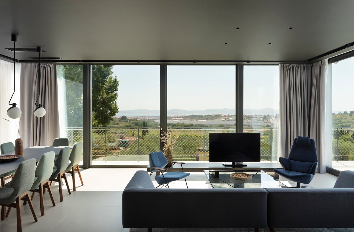 A spacious living area features large glass windows that offer sweeping views of the surrounding landscape. A modern gray sofa is positioned in front of a sleek television, complemented by a stylish blue chair and a light wood dining table with multicolored chairs.
