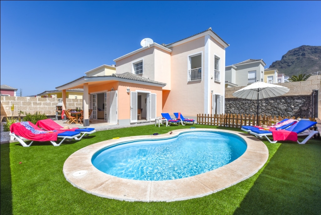 A spacious villa is shown with a well-maintained garden and a private pool, featuring a distinctive shape. Sun loungers are arranged around the pool area, complemented by vibrant beach towels. The exterior walls are painted in soft colors, and clear blue skies provide a bright backdrop.