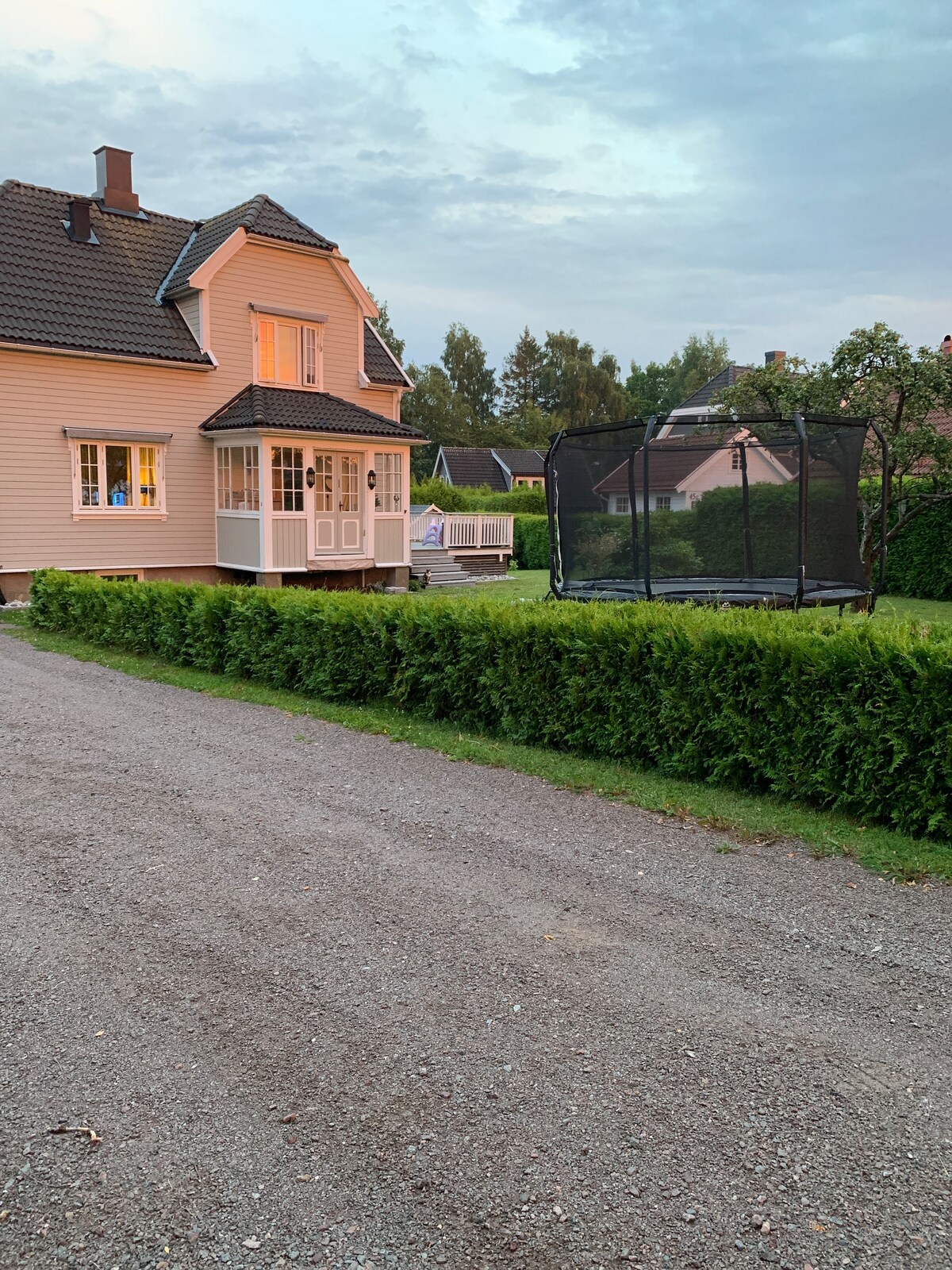 A spacious family home is surrounded by a well-kept garden. The exterior features soft pink siding and large windows, with a trampoline situated on the lawn. A gravel pathway leads to the inviting entrance, framed by neatly trimmed hedges.