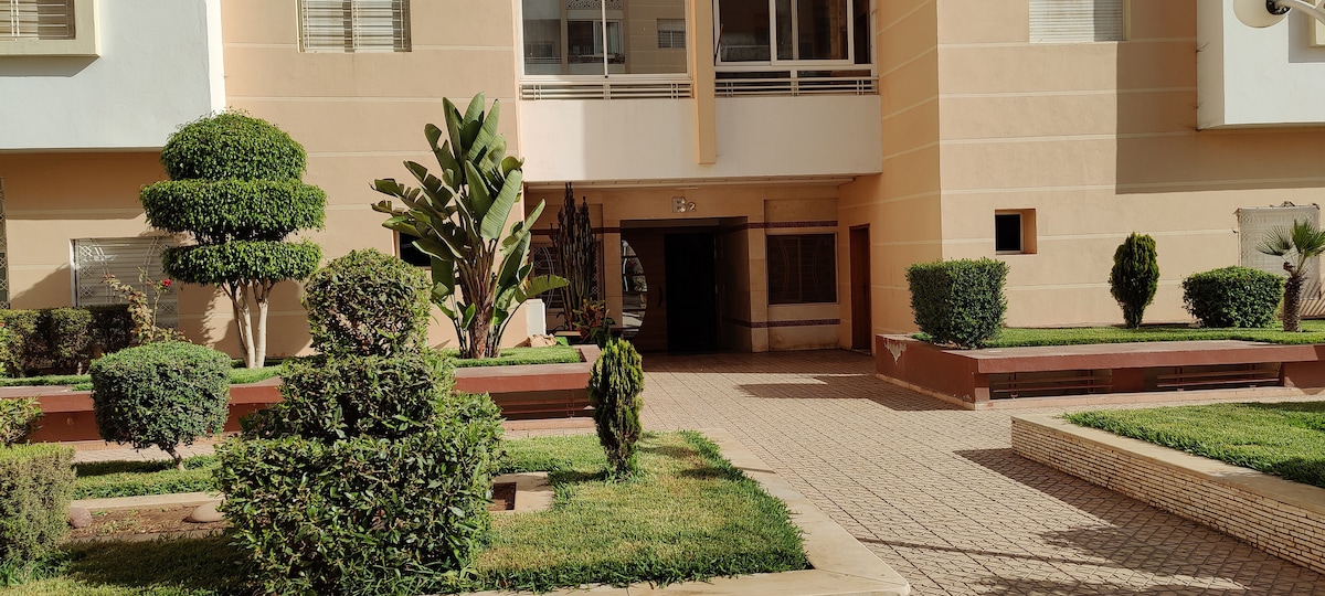A well-maintained exterior courtyard is featured, showcasing manicured greenery and low hedges. Pathways made of light-colored stone lead towards the entrance of the building, bordered by neat garden beds. The building's light-colored facade provides a welcoming backdrop to the landscaping.