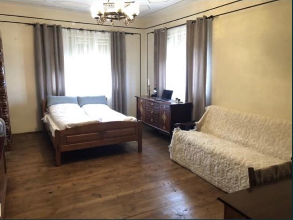 A spacious sleeping area features a light wooden bed with an unmade mattress and soft blue pillows. A light-colored sofa, covered with a textured throw, complements a wooden dresser positioned against the wall. Natural light enters through two partially draped windows.