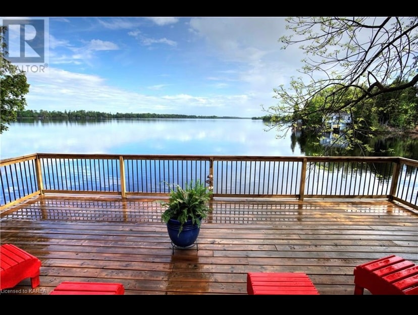 A spacious wooden deck overlooks the calm waters of Beaver Lake, framed by lush greenery. Red outdoor chairs provide comfortable seating, while a blue planter adds a touch of color. The scene is serene, reflecting the tranquility of the surrounding nature.