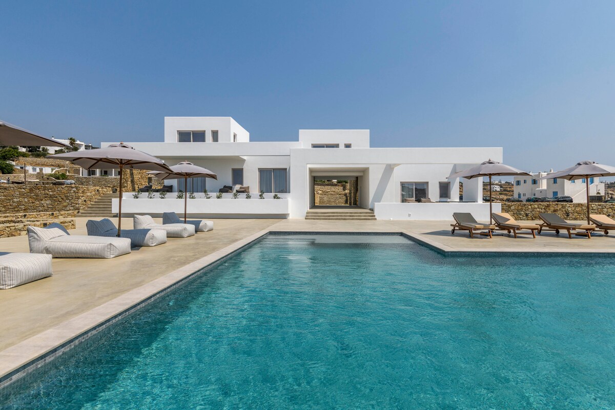 A modern building with a clean white facade is set against a clear blue sky. Lounge chairs and umbrellas surround a spacious pool, creating a relaxed outdoor area that opens up towards the entrance of the suite. The layout highlights the tranquil atmosphere.