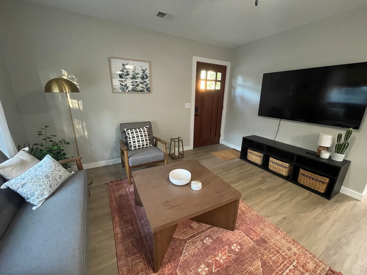 The living area features a modern design, with a comfortable gray sofa and two accent chairs. A wooden coffee table is centered on a patterned area rug. A flat-screen TV is mounted on the wall, and natural light enters through the front door's glass panels.