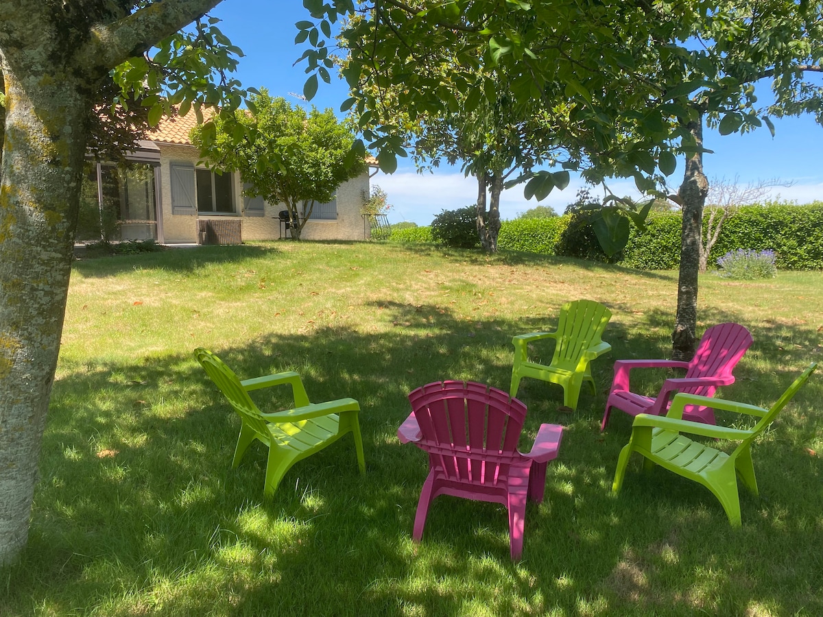 A grassy area is depicted, featuring five colourful chairs arranged around a small gathering space. Shade is provided by nearby trees, and a house is visible in the background, surrounded by lush greenery.