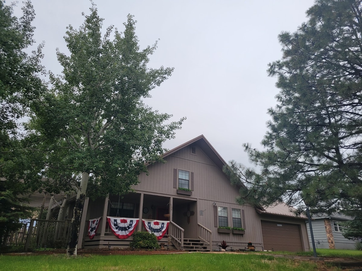 A two-story home with a light brown exterior is set among lush trees. A welcoming porch is decorated with patriotic bunting. Greenery surrounds the house, emphasizing the outdoor space. The structure features a sloped roof and a mix of vertical and horizontal siding.