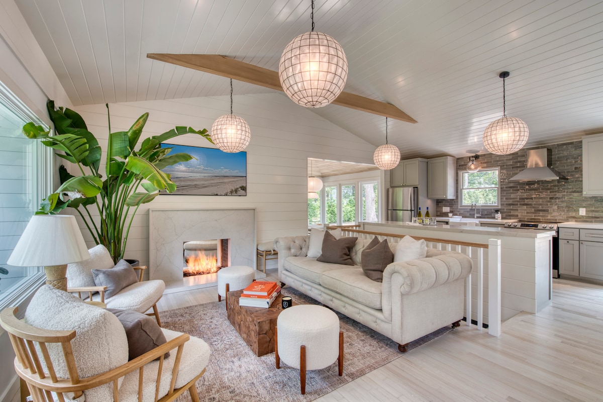 A bright living space features a classic sofa, two armchairs, and decorative lighting. A stone fireplace adds a cozy element, while a large window invites natural light. Indoor plants provide greenery, and a kitchen area with modern appliances is visible in the background.