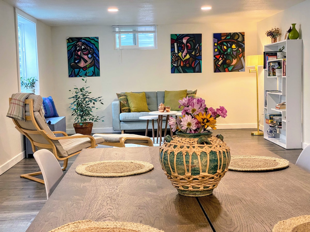 The spacious living/dining area features a wooden dining table set with woven placemats, surrounded by white chairs. A light-colored sofa, adorned with bright cushions, is positioned nearby. Colorful artwork decorates the walls, and a potted plant adds a touch of greenery to the space.