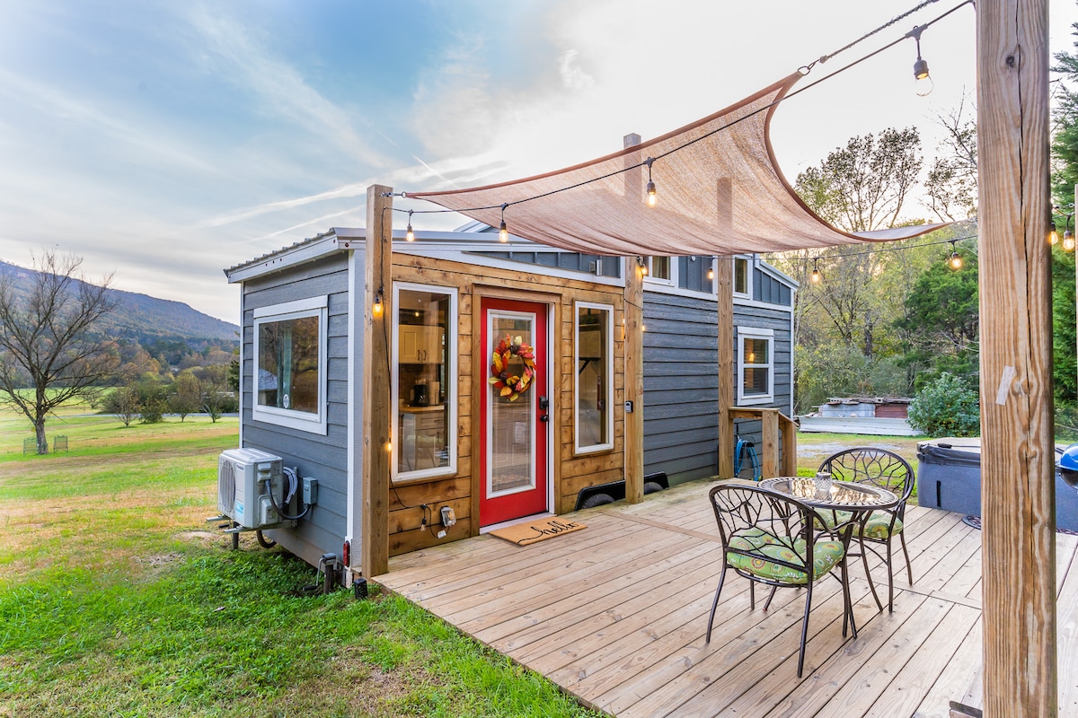 A charming exterior of the tiny home is showcased, featuring a red front door adorned with a wreath. A wooden deck with a small dining table and metal chairs is visible, complemented by string lights overhead and surrounded by lush grassy meadows and mountain views.