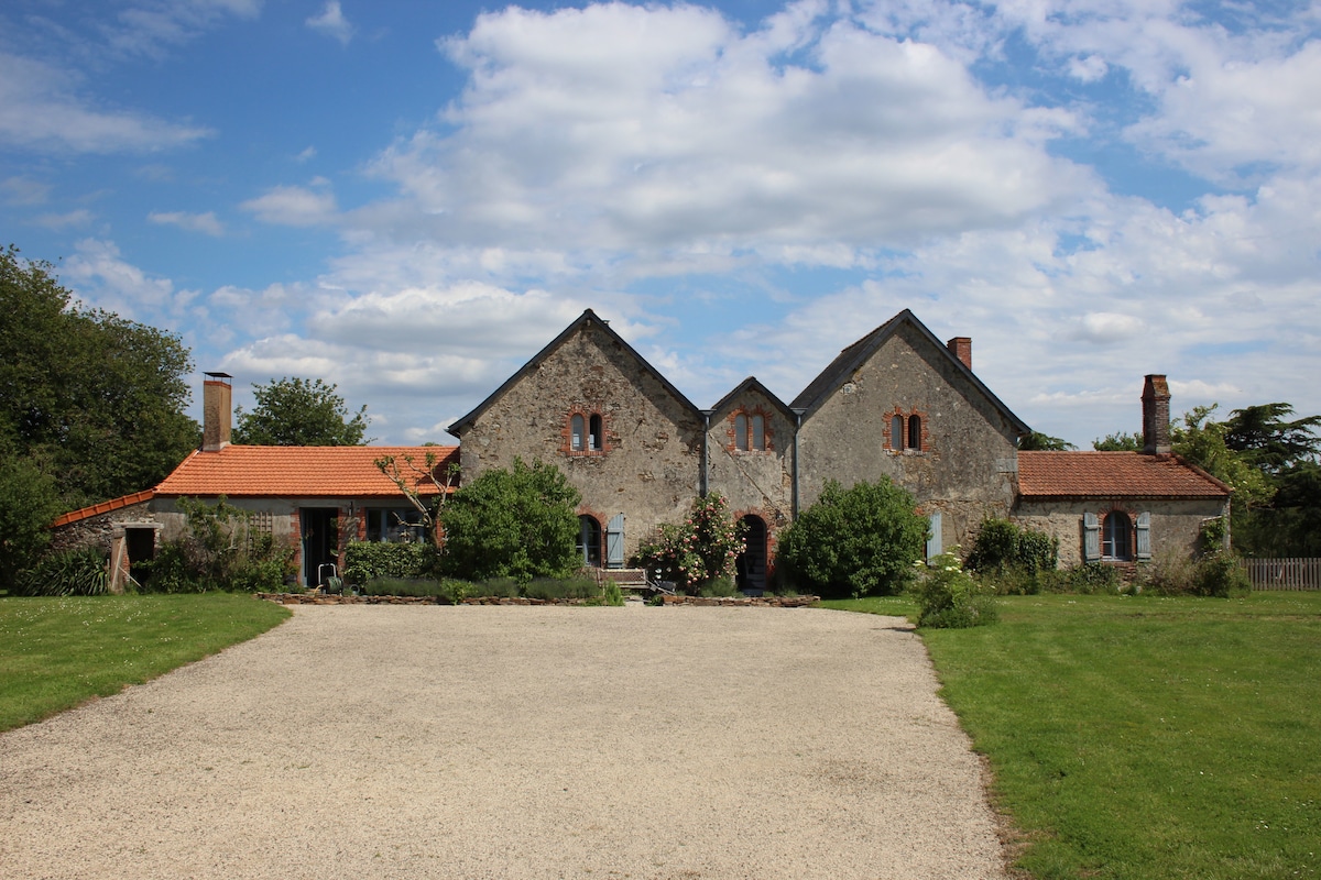 A charming stone house with a rustic façade is set amidst a green landscape. The building features distinct roofs and several windows. A gravel path leads to the entrance, flanked by landscaped gardens, enhancing the serene countryside ambiance.