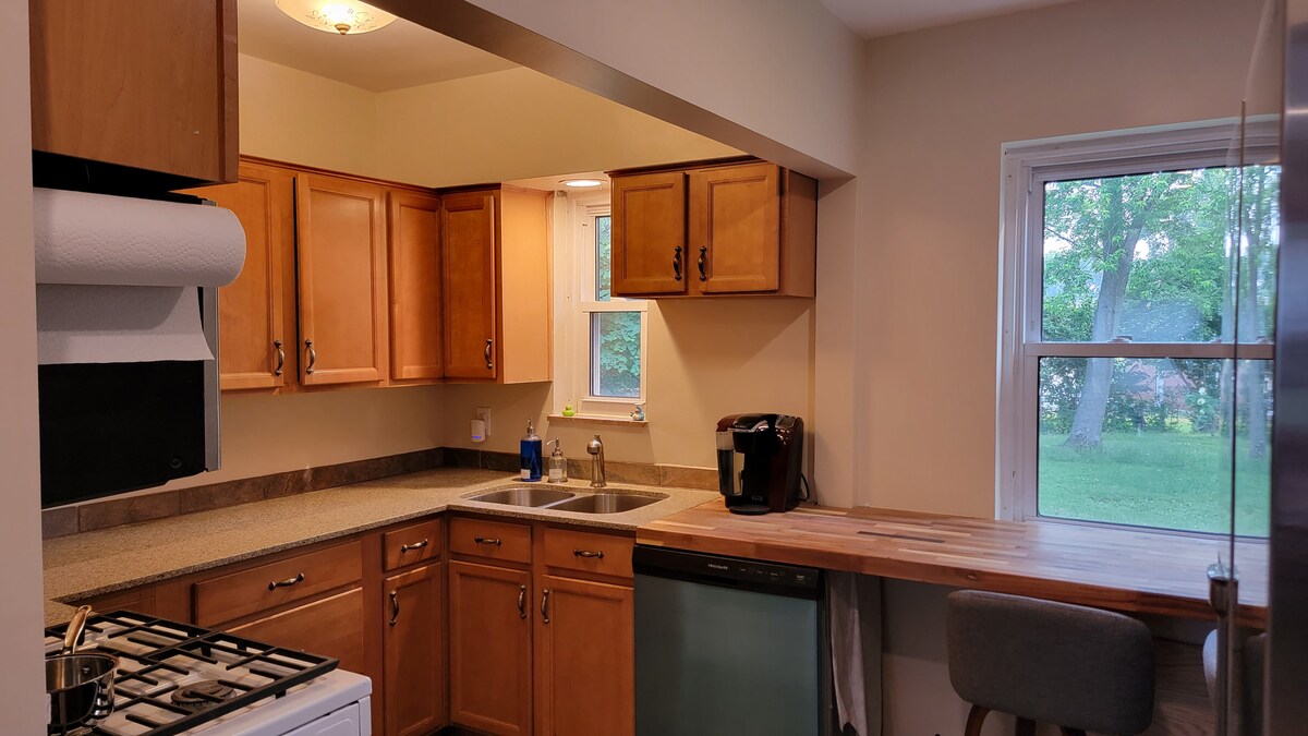 The kitchen features wooden cabinetry and a large window, allowing natural light to fill the space. A sink is positioned between the stove and a countertop with a bar stool. A coffee maker is visible on the counter, enhancing the room's functionality.