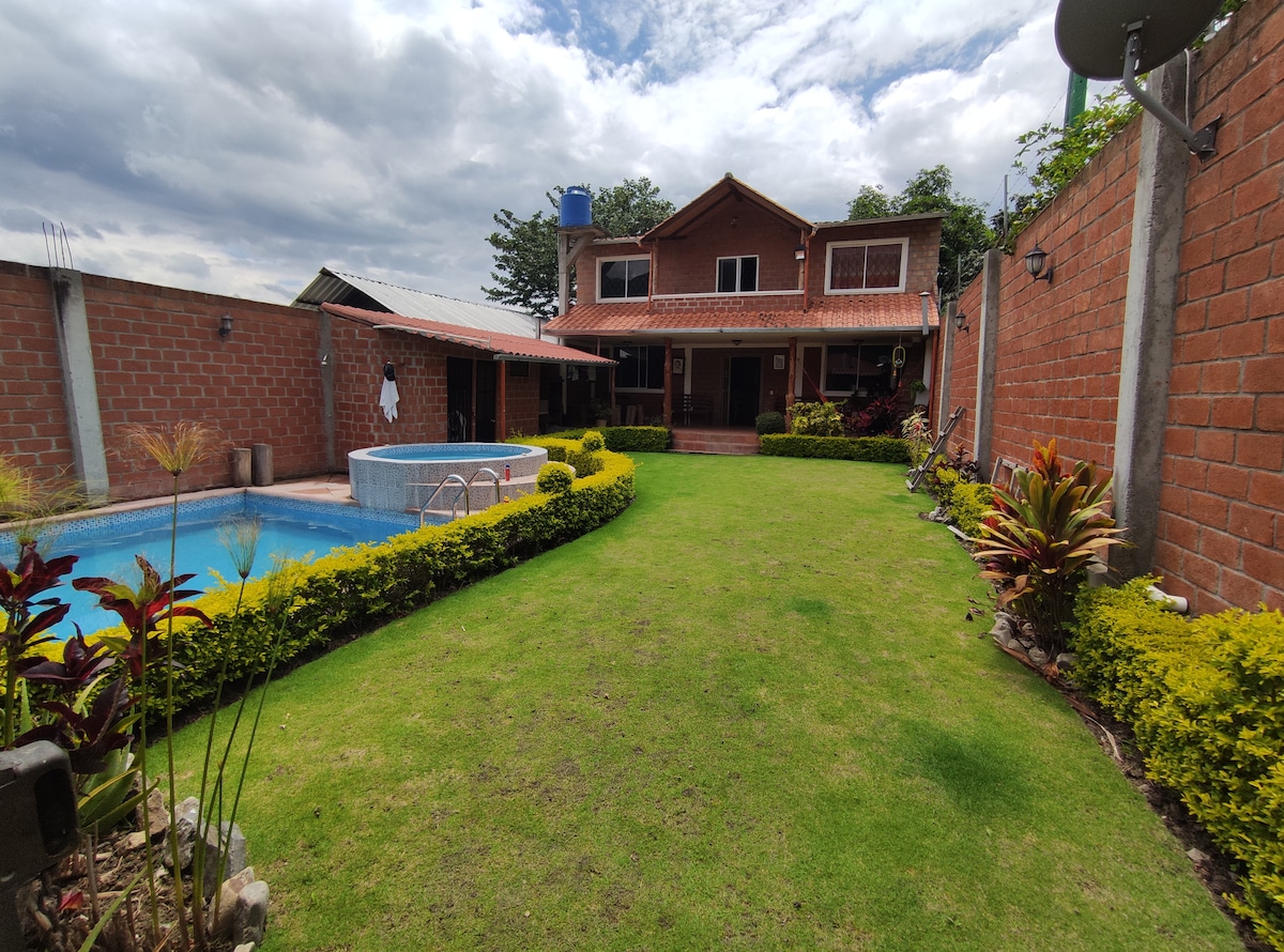 The image displays a two-story vacation house surrounded by well-maintained greenery. A clear swimming pool is visible in the foreground, along with a neatly manicured lawn. The brick exterior of the house is complemented by various plants along the pathways and a patio area.