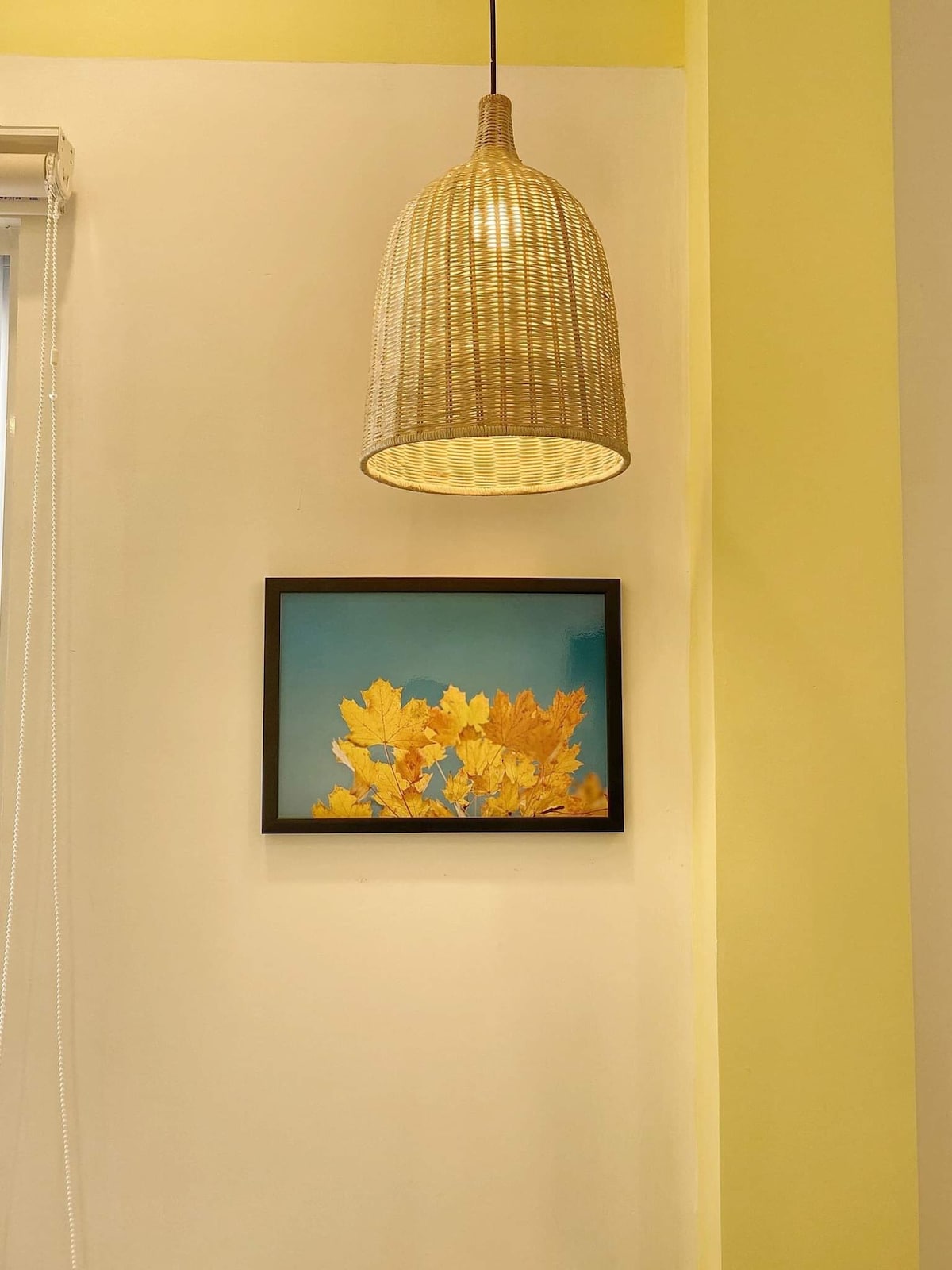 A woven light fixture hangs from the ceiling, casting a warm glow over a framed artwork. The artwork features vibrant yellow leaves set against a bright blue background, providing a cheerful contrast to the soft yellow wall.