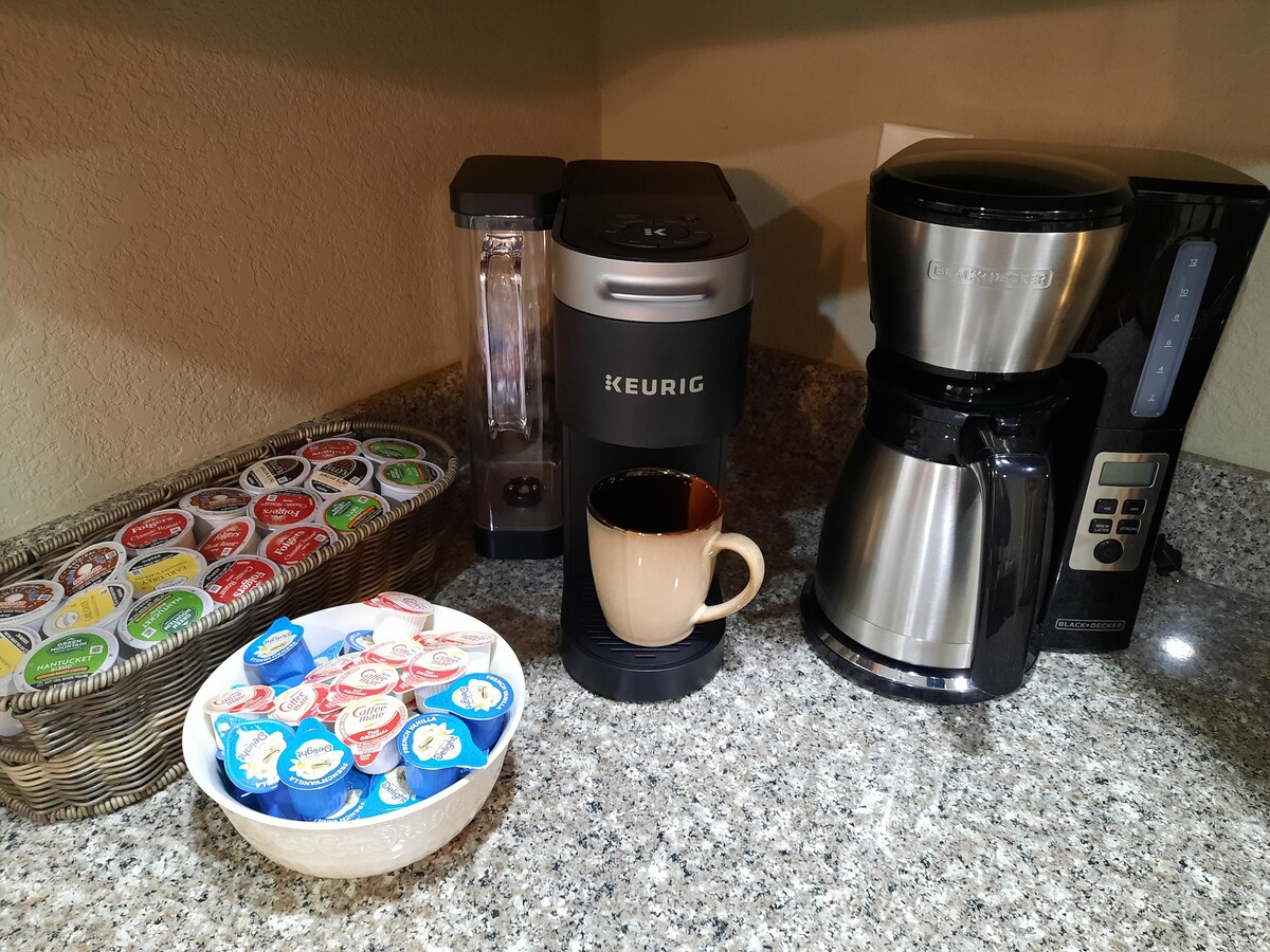 A coffee station is set up with a Keurig machine and a traditional coffee maker on a granite countertop. A basket filled with various coffee pods is displayed alongside a bowl containing creamers. A coffee cup is positioned in front of the Keurig.