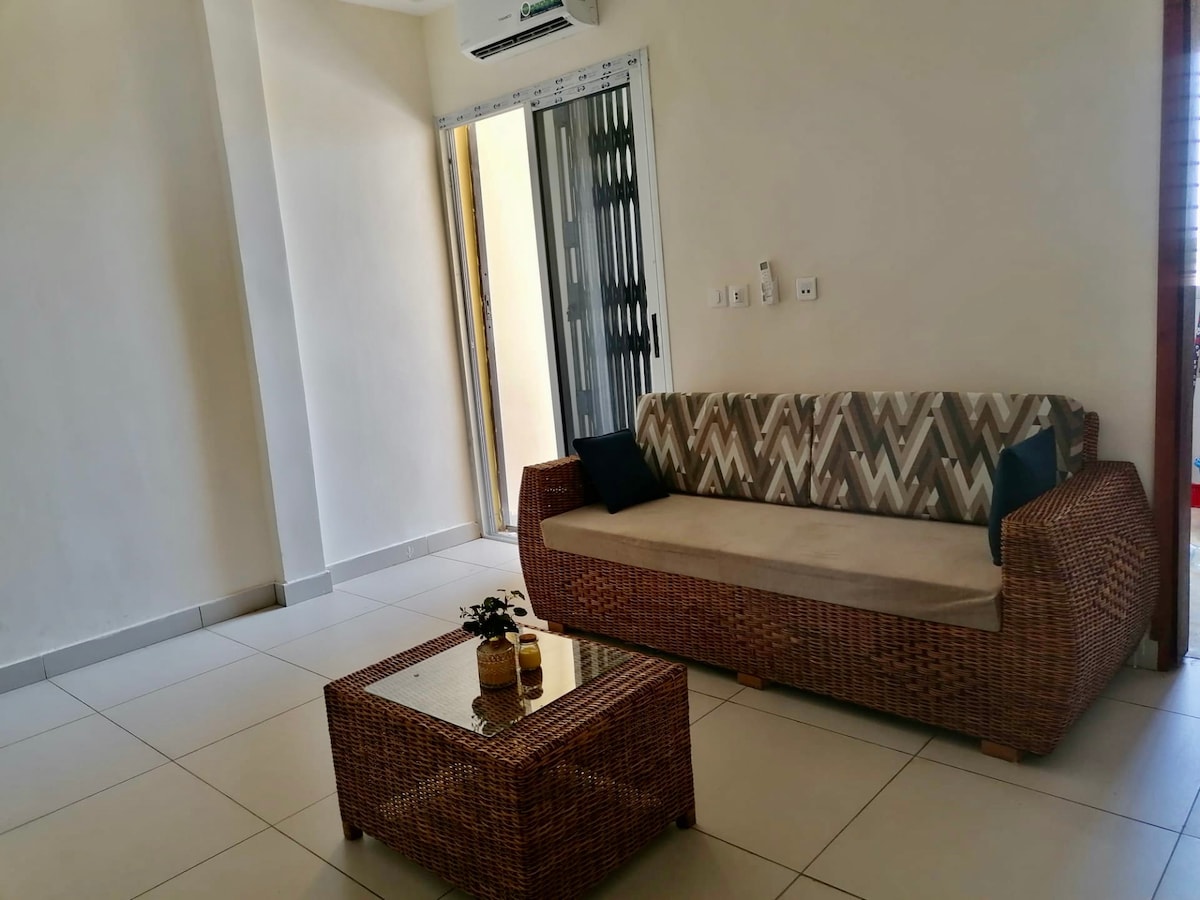 A cozy seating area features a woven sofa adorned with patterned cushions and two dark accent pillows. A glass-topped coffee table sits in front, displaying a small decorative item. Natural light enters through a nearby door leading to an outdoor space.