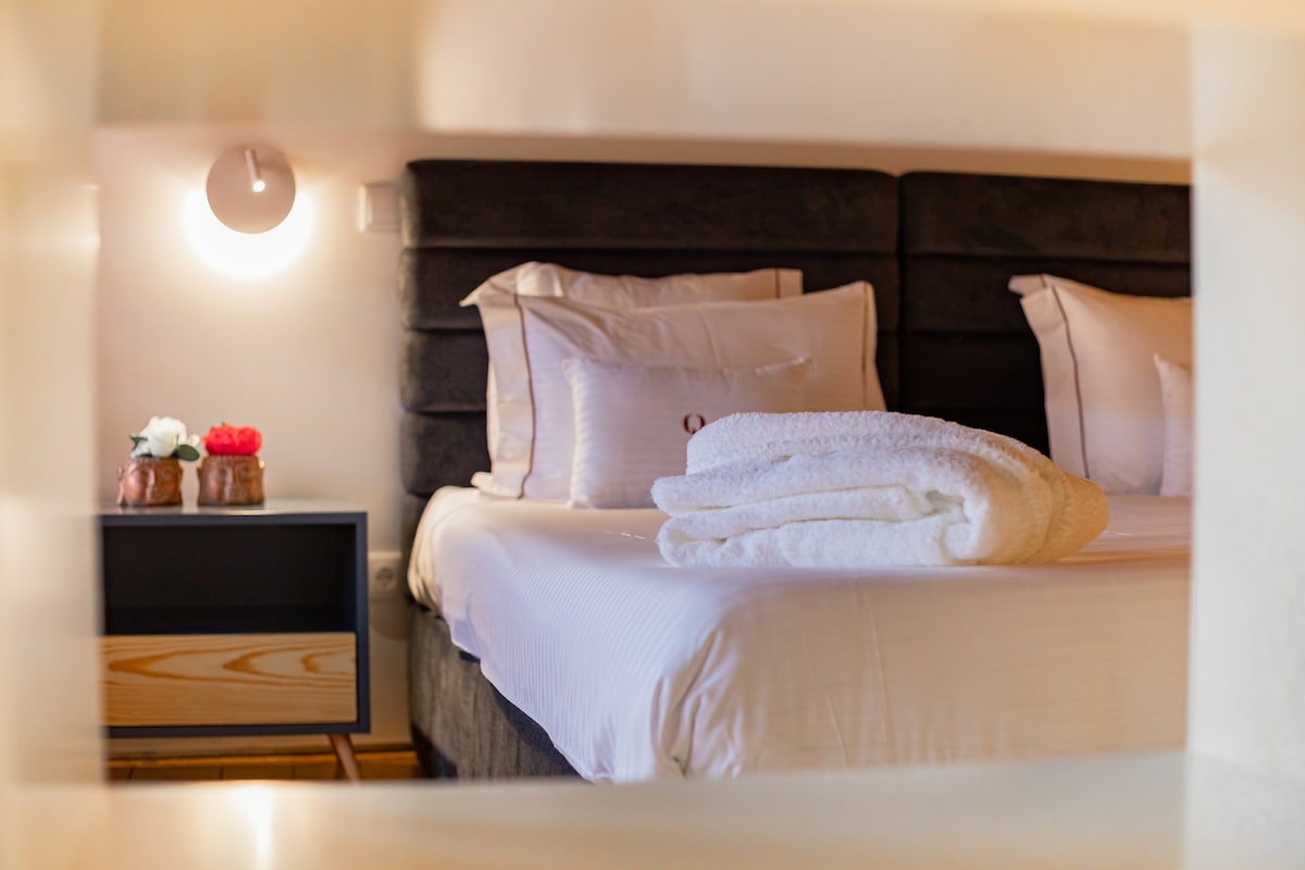 A spacious bed is presented with neatly arranged white towels. Soft lighting from a wall lamp highlights the headboard, while a small nightstand is adorned with a vase of flowers, adding a touch of elegance to the room.