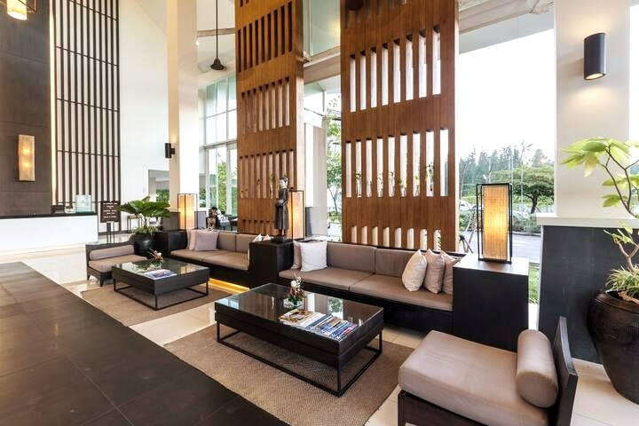 A modern lobby area is showcased, featuring dark wooden accents and a high ceiling. Comfortable seating is arranged around a low table, and natural light flows through large windows. Stylish plants add a touch of greenery to the inviting space.